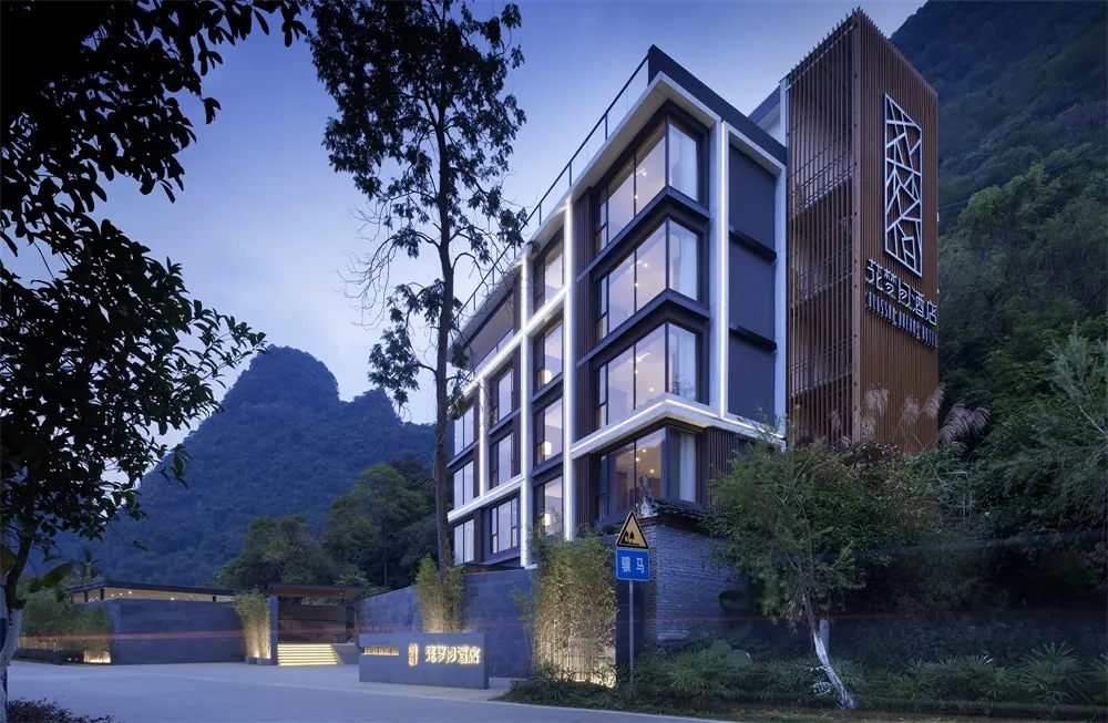 A modern, multi-story resort building with large windows and wooden accents, surrounded by lush greenery and mountains in the