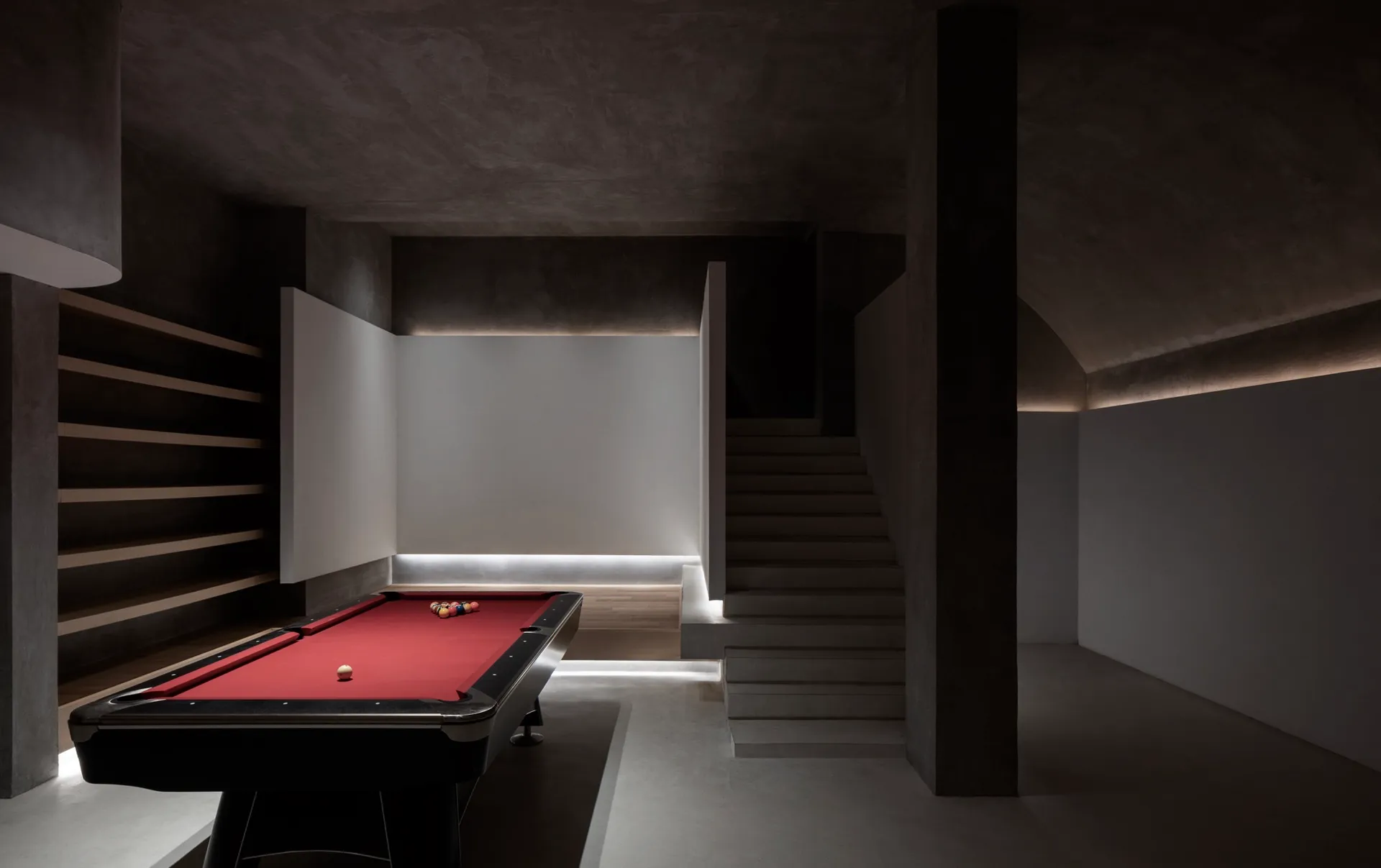 A minimalist industrial game room featuring a red pool table, concrete walls, and a lamp.