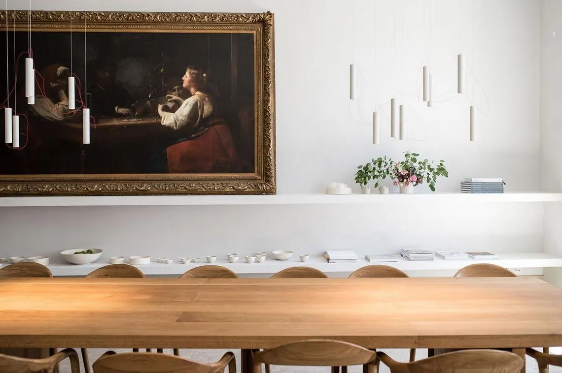 A minimalist dining room with a wooden table and chairs, featuring a hanging light fixture and a framed painting on the wall.