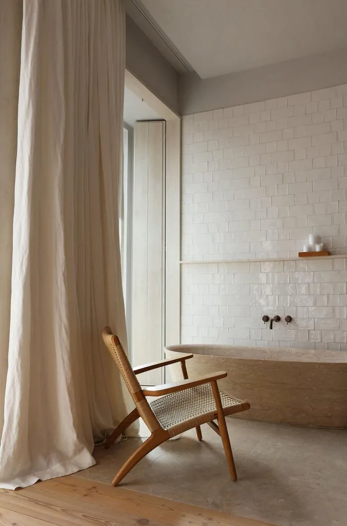 A modern bathroom featuring a tile wall and concrete floor, with a wooden armchair in the foreground and a large shower curta