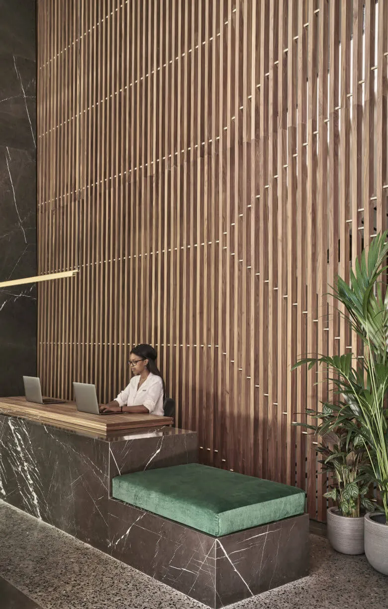 A modern lobby interior featuring a marble desk with a green cushion and a wooden grille wall behind it.