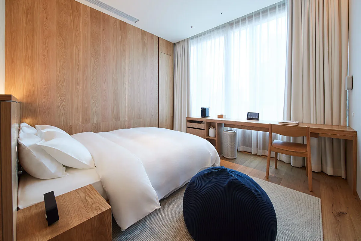 A modern minimalist bedroom featuring wood walls, a white bedspread, and a navy pouf.