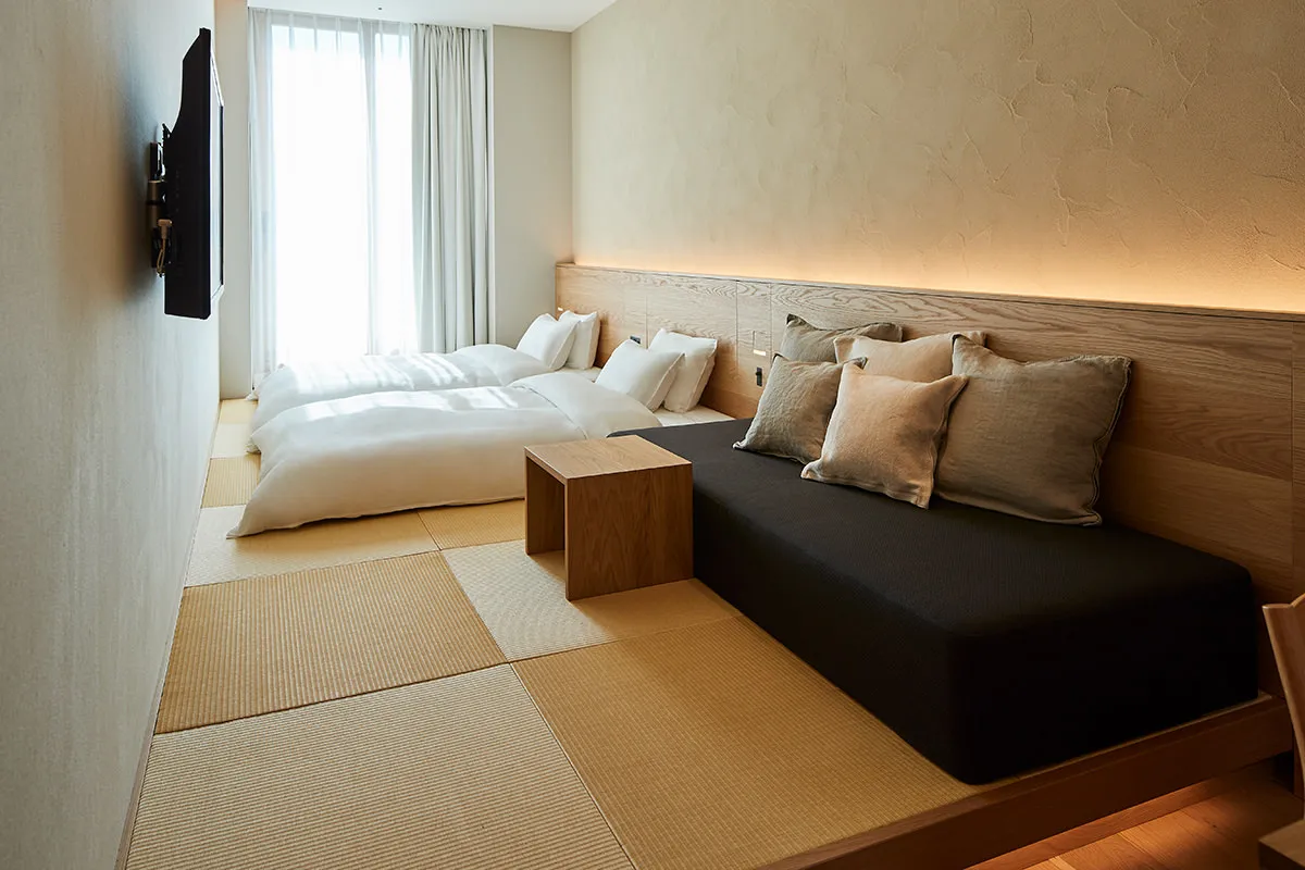 A minimalist bedroom featuring a wood-framed bed and a black sofa with cushions, set against a pale neutral background with s