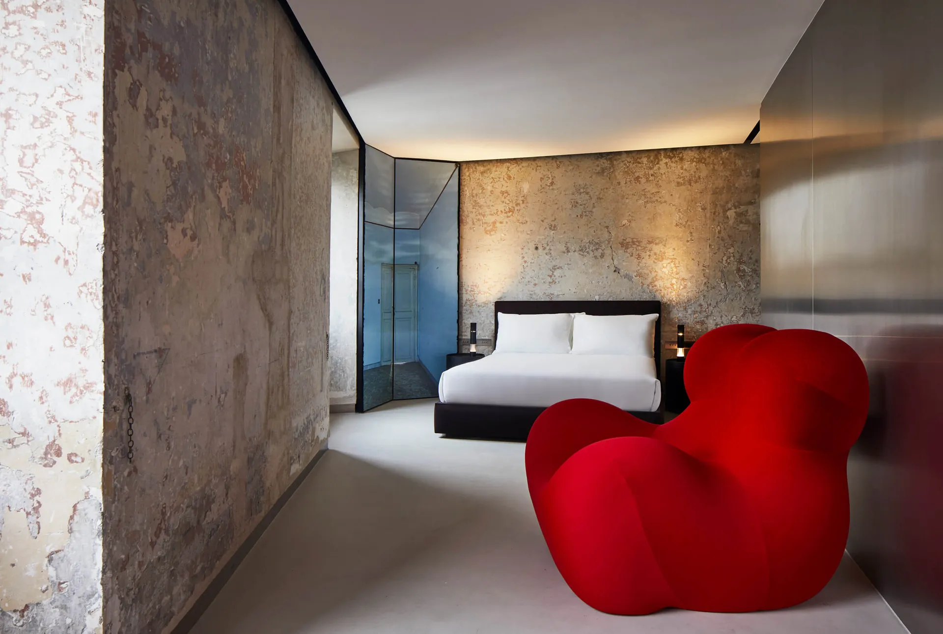 Modern bedroom featuring a concrete wall, a red chair, and a neatly made bed with white sheets and pillows.