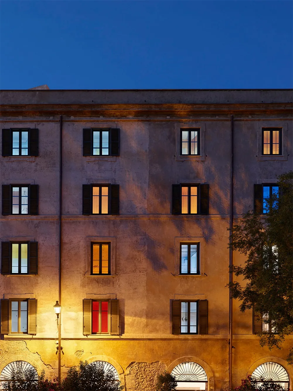 A stone building facade illuminated at dusk with lights on inside several windows.