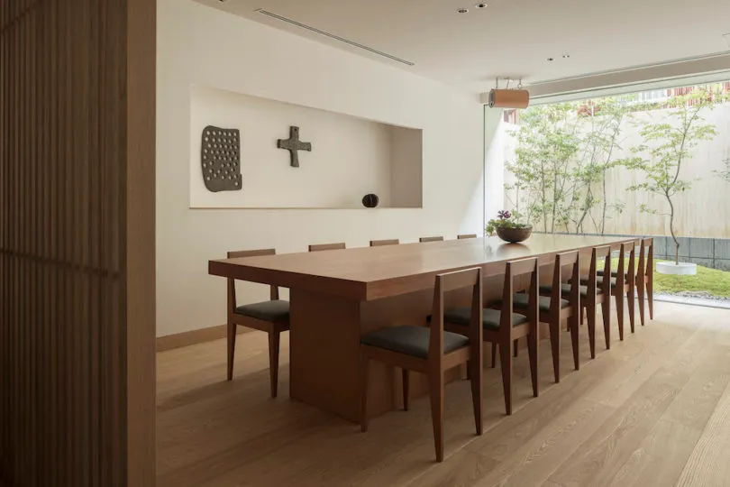 A minimalist dining room featuring a long wooden table surrounded by chairs, with a minimalist wall art piece and a small gar