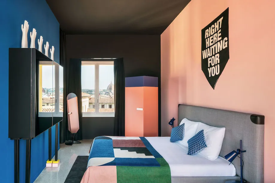 A modern bedroom with pink and blue walls, a patterned blanket, and a poster reading 'Right Here Waiting for You'.