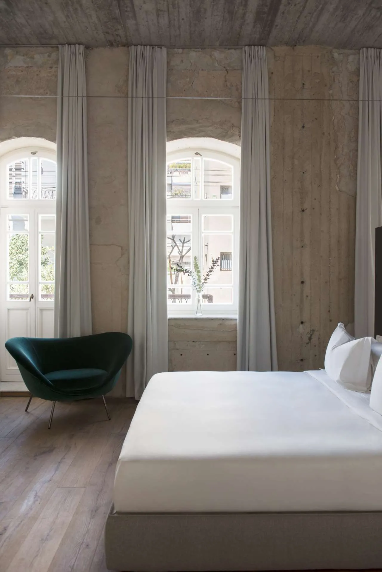 A minimalist bedroom featuring a white bed with white linens, a green armchair, and white curtains.