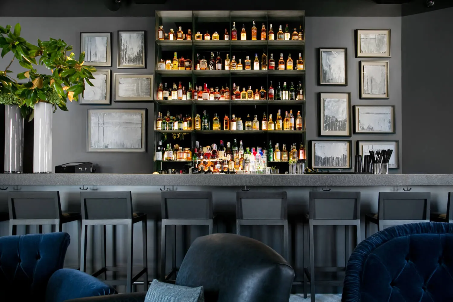 Modern bar interior with sleek metallic shelving, glass bottles, leather seating, stone countertops, and wooden accents.