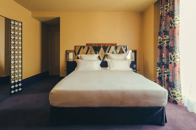 A spacious modern bedroom featuring a bed with white linens, a geometric headboard, and colorful curtains against a mustard y