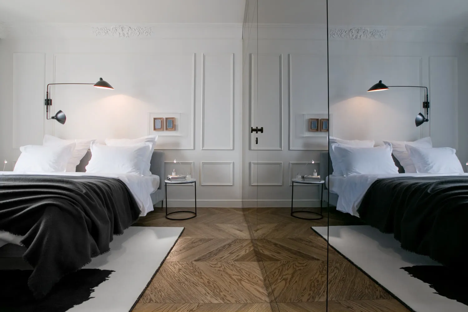 A minimalist bedroom with a wood floor and white walls, featuring a bed with white bedding and a dark throw blanket, illumina