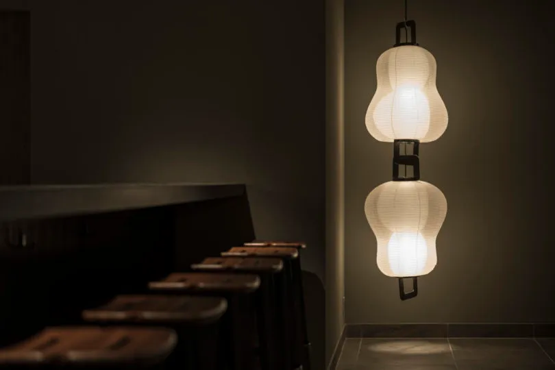 A row of stools in a dimly lit restaurant bar with a minimalist design, featuring a decorative paper lamp hanging from the ce