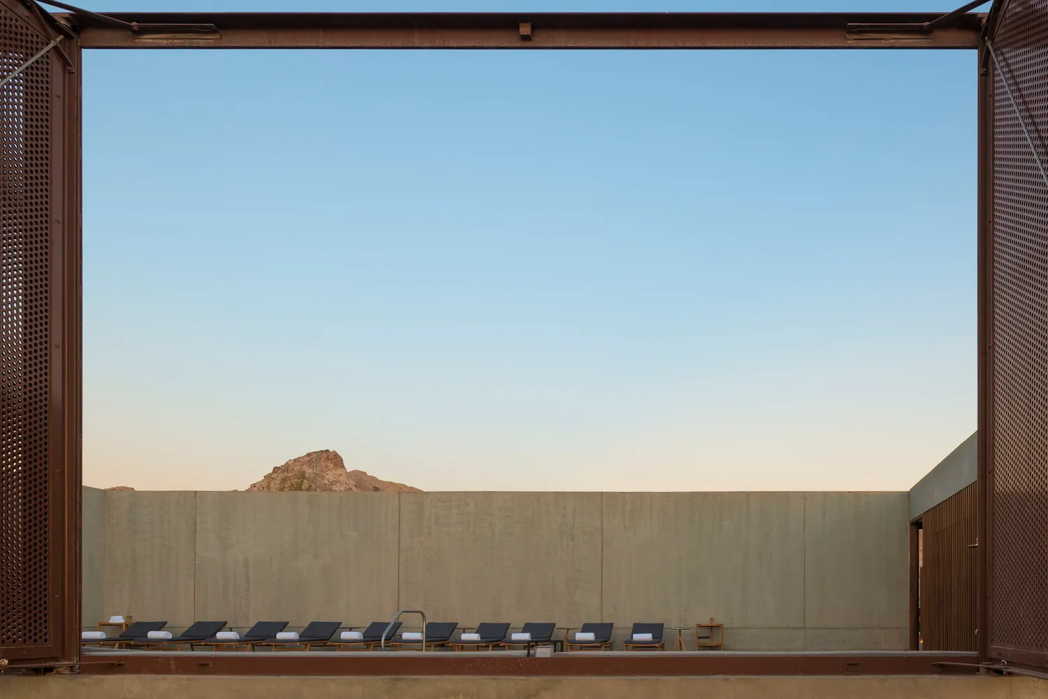 A minimalist poolside area with concrete walls, featuring several lounge chairs lined up under a clear blue sky.