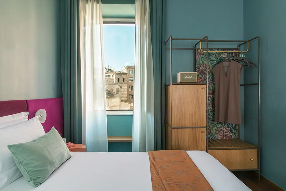 Modern hotel room with neutral bed and furniture, blue walls, and a view outside through sheer curtains.