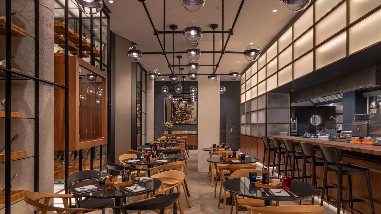 A modern industrial restaurant interior with sleek black pendant lights, wooden tables, and glass railings.