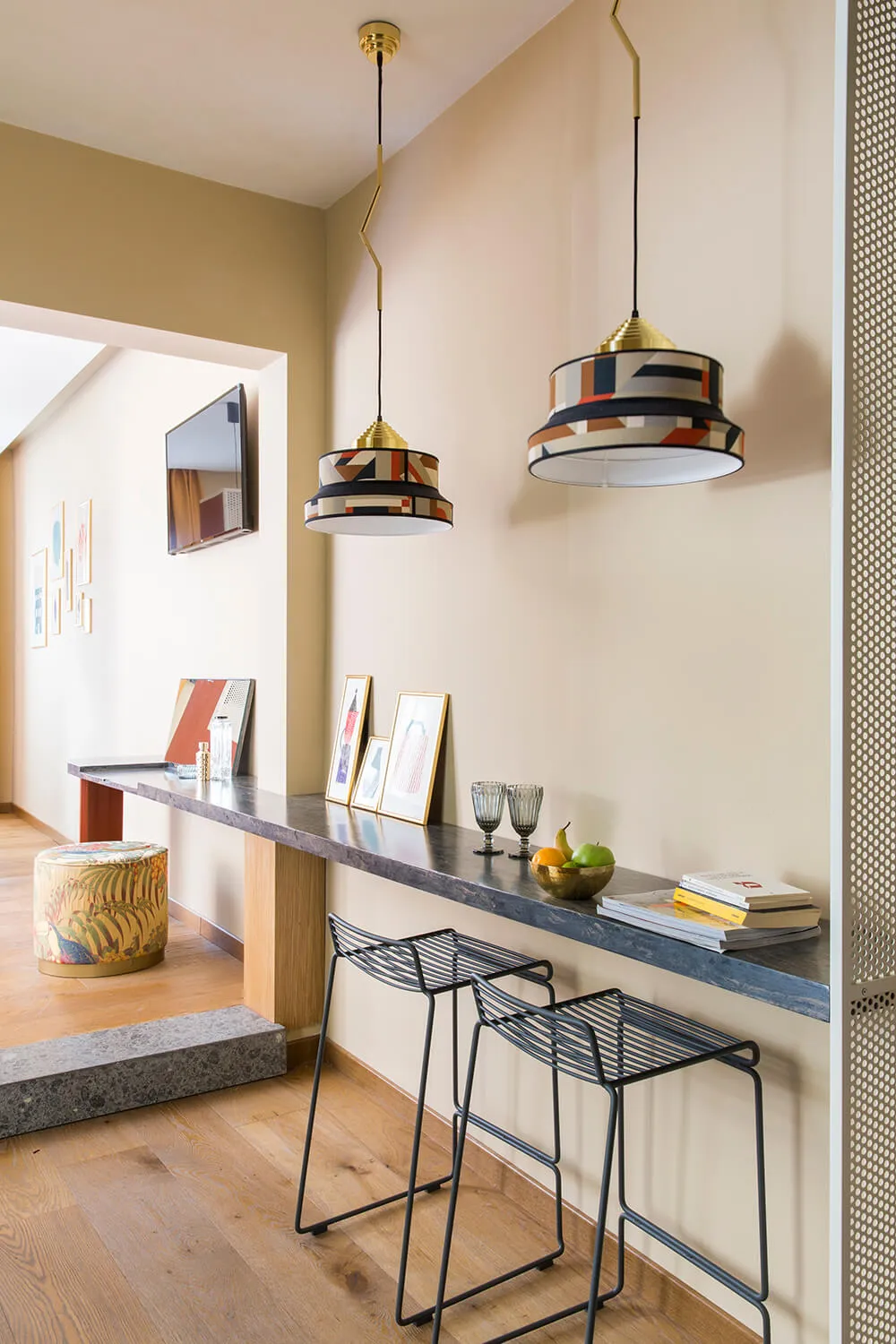Modern kitchen with marble countertops, colorful pendant lights, and black stools. Wooden floor and framed art on the wall.