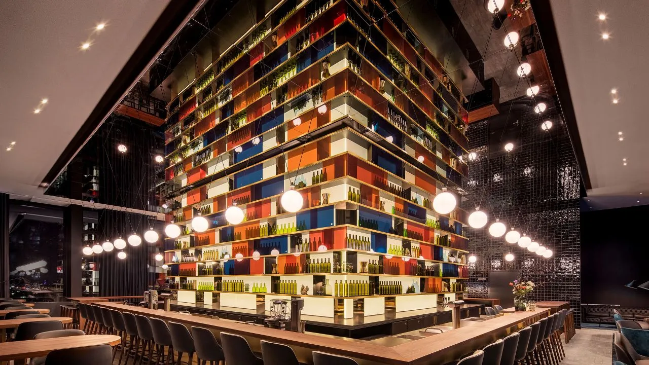 A modern restaurant interior with wooden tables and chairs, colorful shelves, and hanging lights.