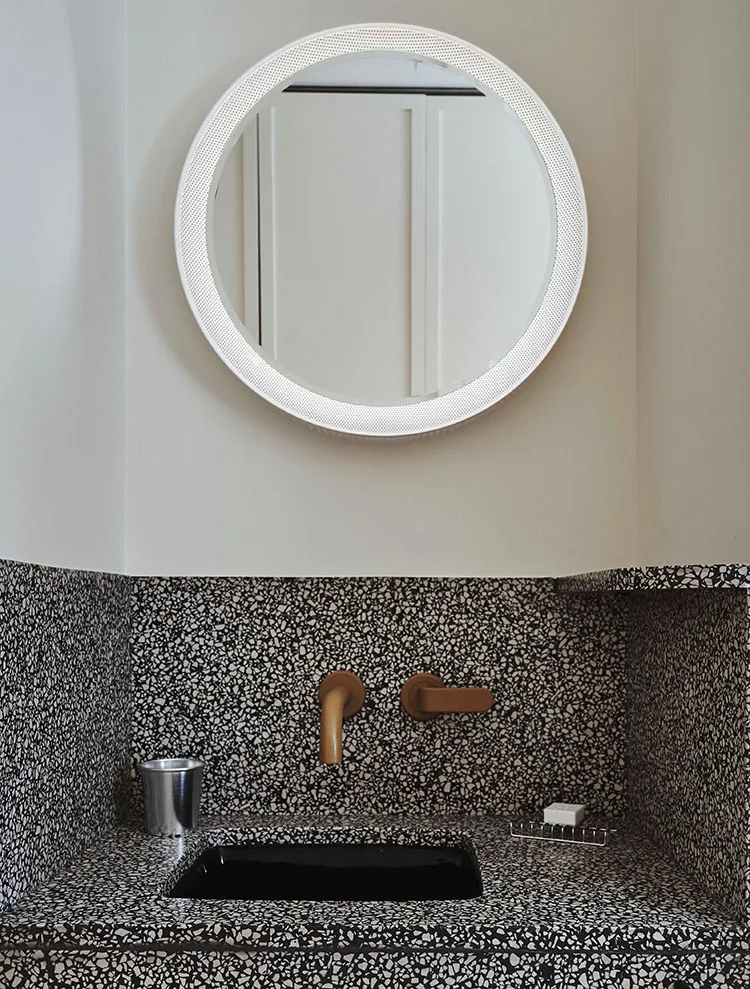 A modern bathroom with a black and white terrazzo countertop, a black porcelain sink, and a round mirror with a perforated fr