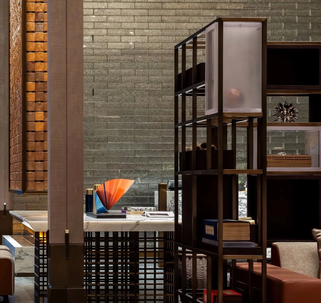 A modern lobby interior with metallic accents and glass elements, featuring a contemporary art piece and shelving unit.