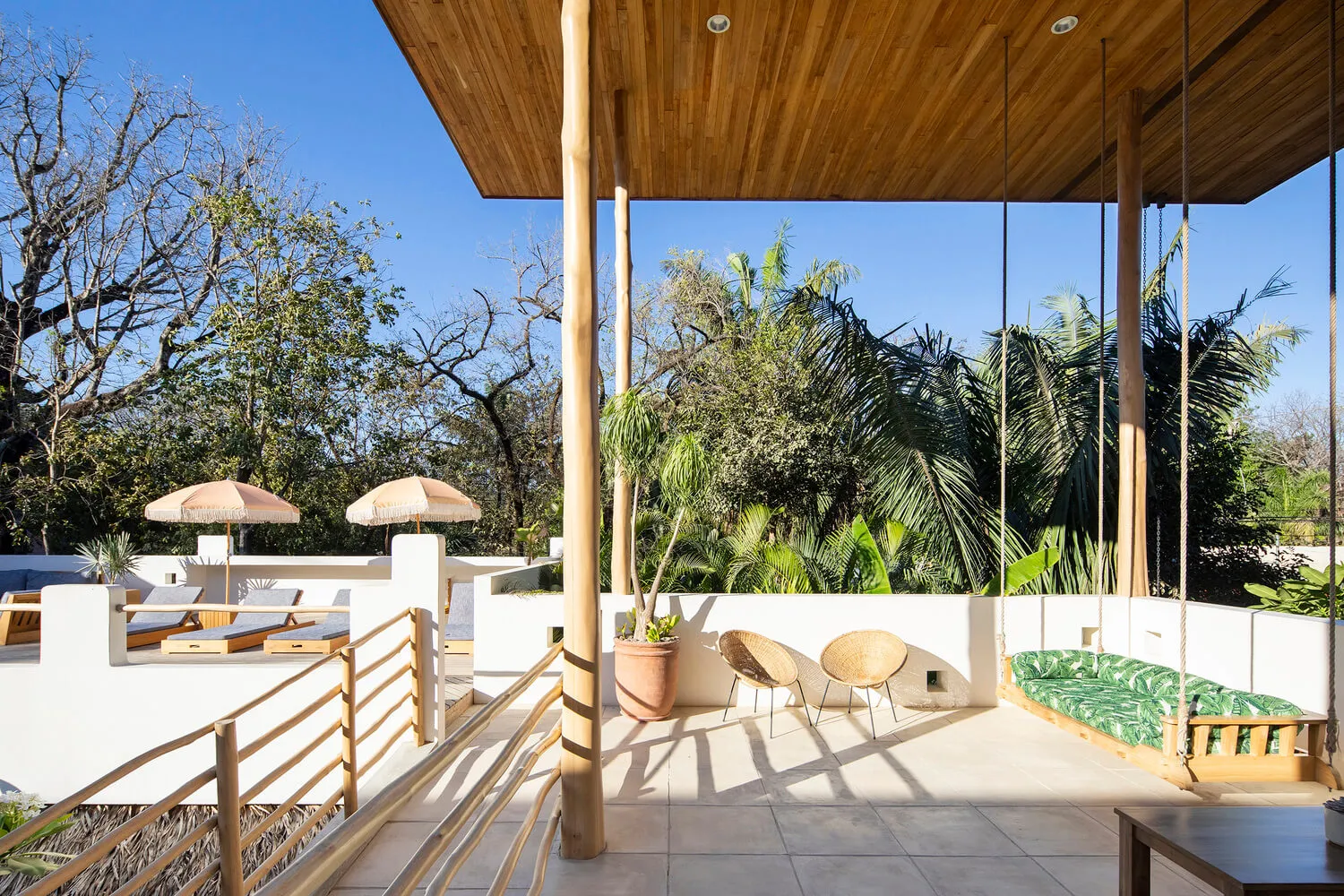 An outdoor wooden patio surrounded by tropical plants and featuring a hanging bench and lounge chairs under a wooden roof.