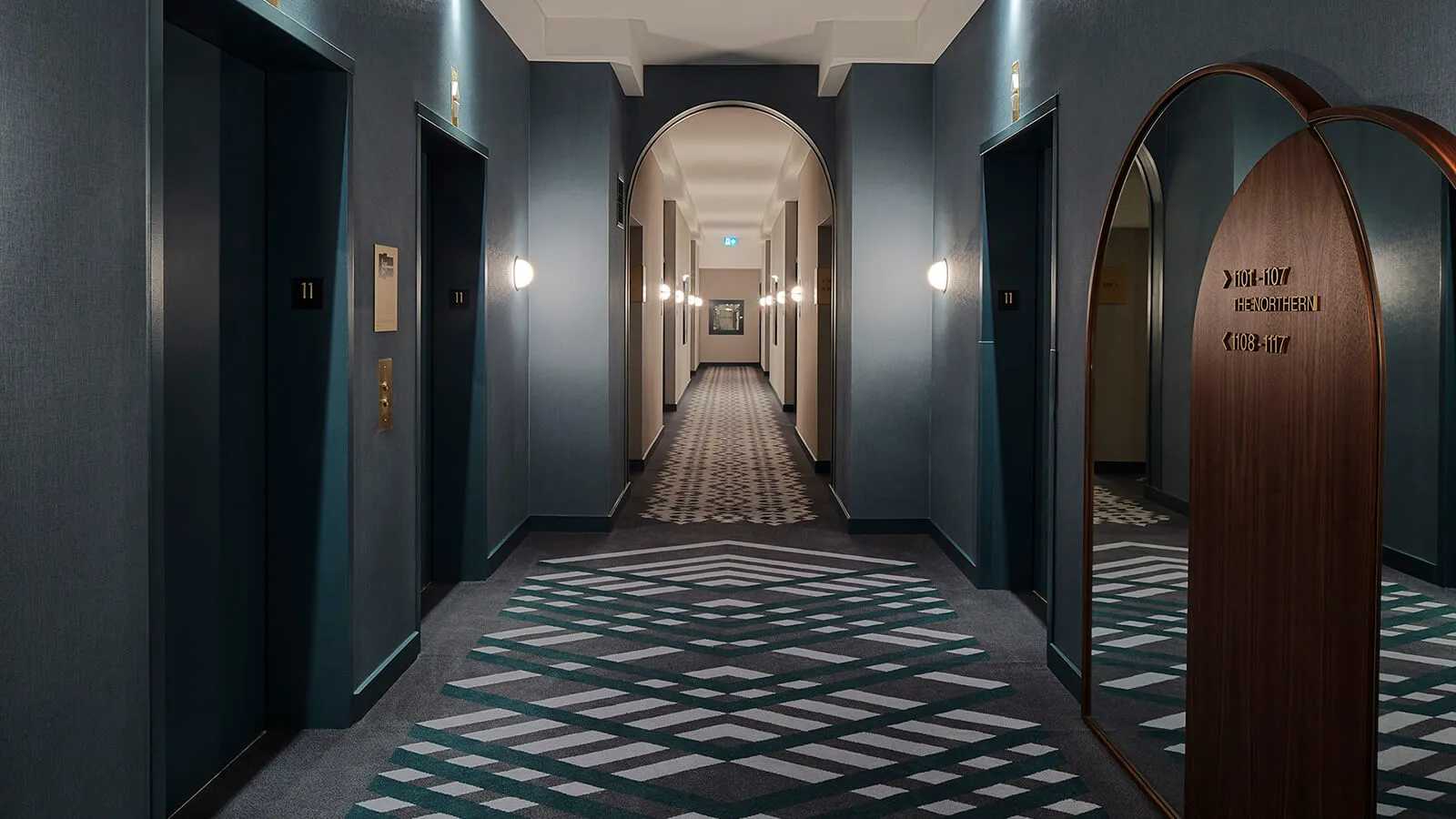 A modern hotel hallway featuring a patterned carpet and wooden accents.