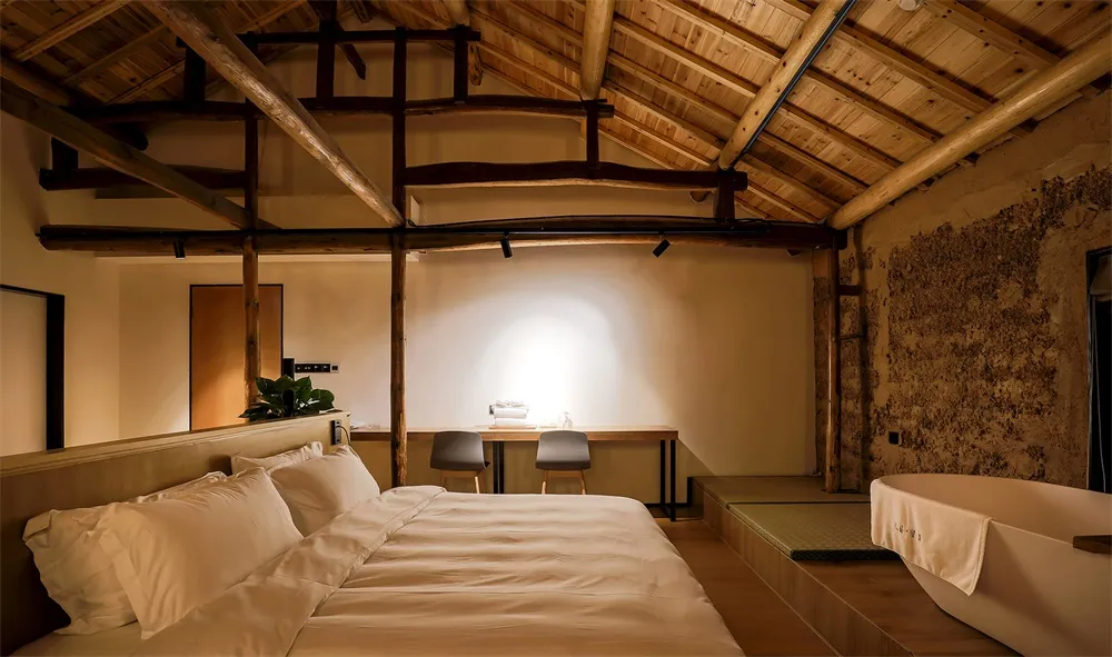 A minimalist bedroom featuring wooden beams on the ceiling, a bed with white bedding, and a small desk area.