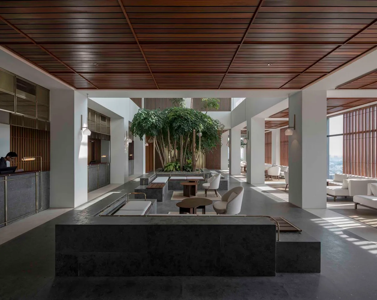 Lobby area of a modern hotel with concrete benches, white chairs, and a central greenery feature.