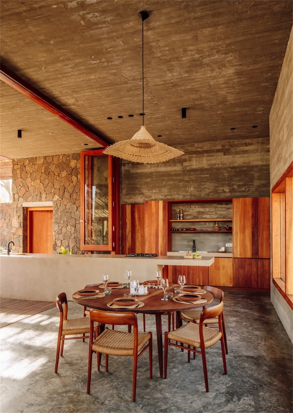 A modern dining room featuring a round wooden table, chairs with woven seats, concrete floor, stone wall, and exposed wooden