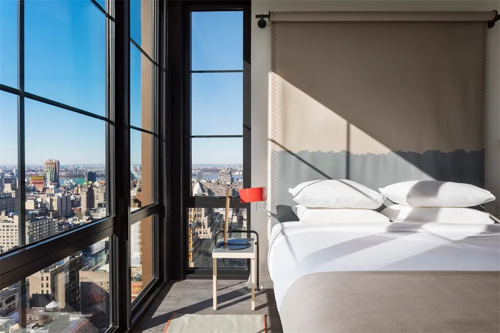A modern glass bedroom with a large bed and cityscape view through large windows.