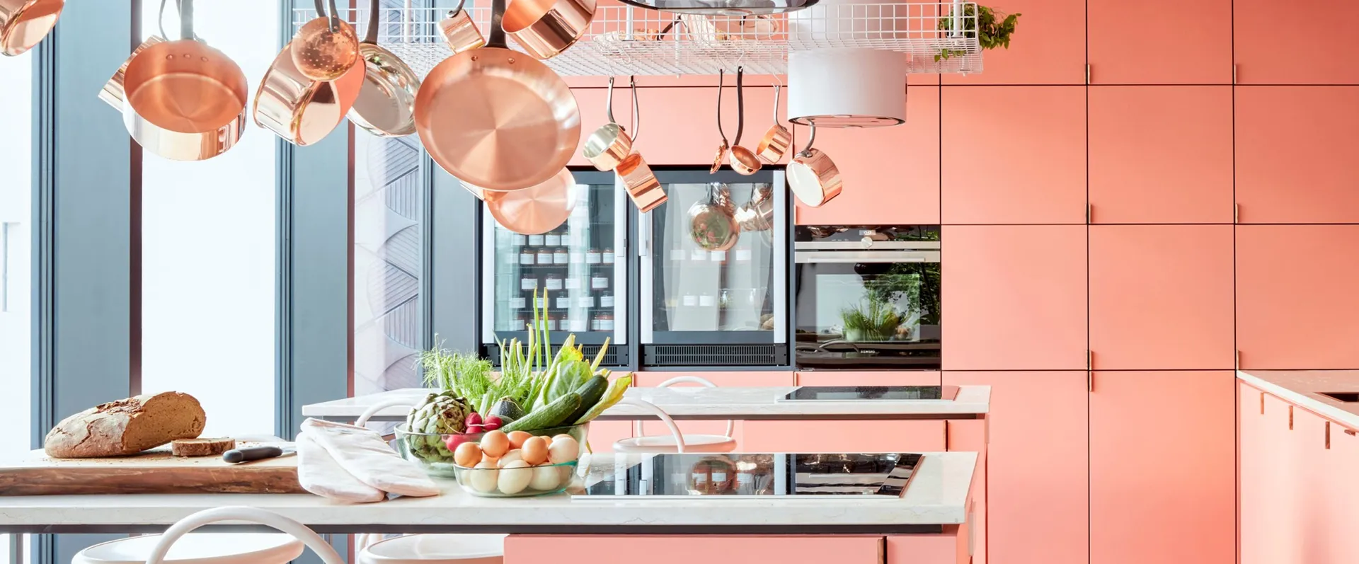 A modern, minimalist kitchen with pink cabinets, copper pots and pans hanging overhead, and a variety of fresh produce on the