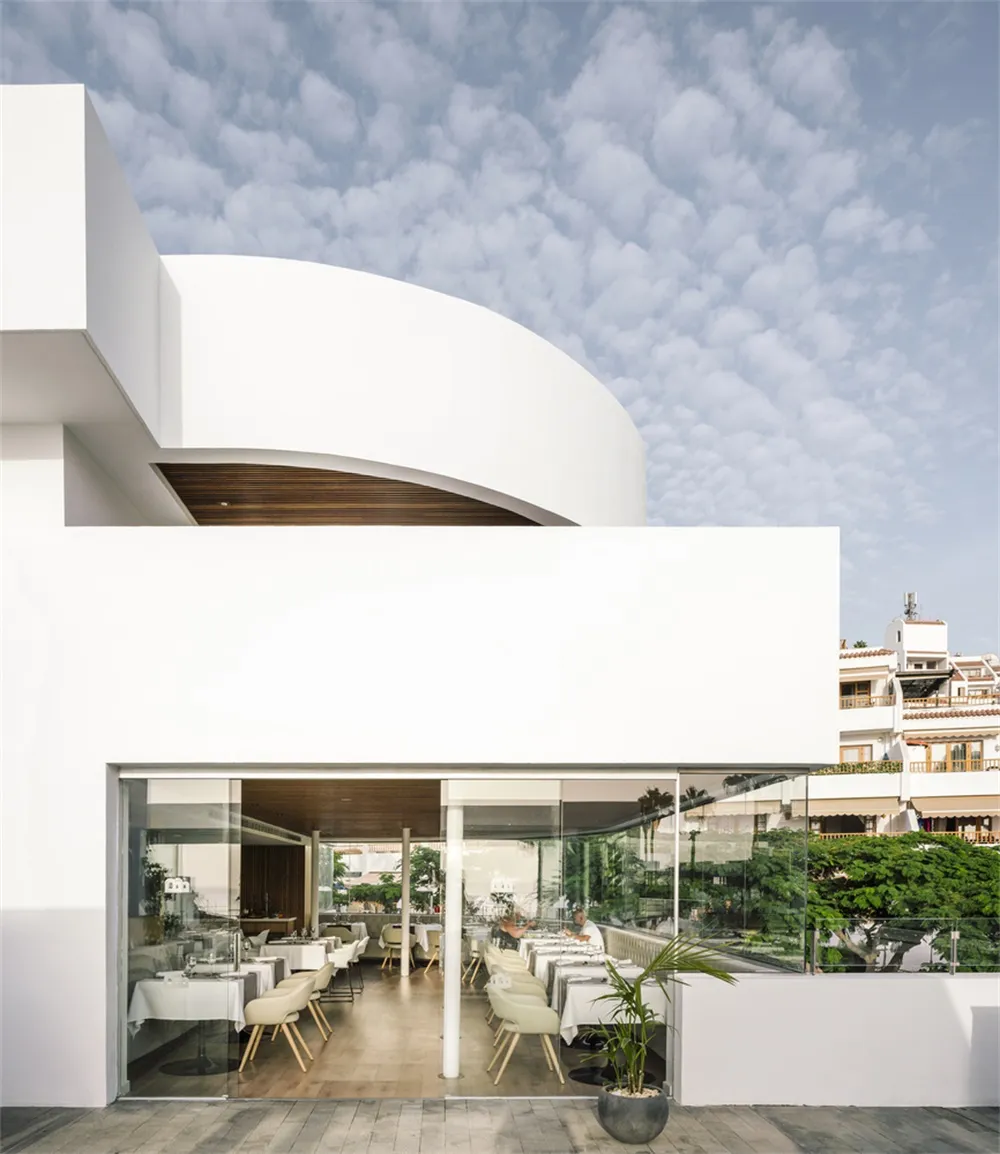 White minimalist restaurant with glass walls and white furniture under a cloudy sky.