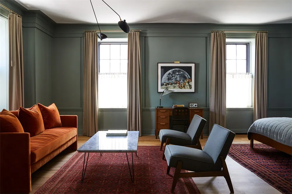 A modern living room with an orange velvet sofa, two gray armchairs, a wooden desk, and a red rug.