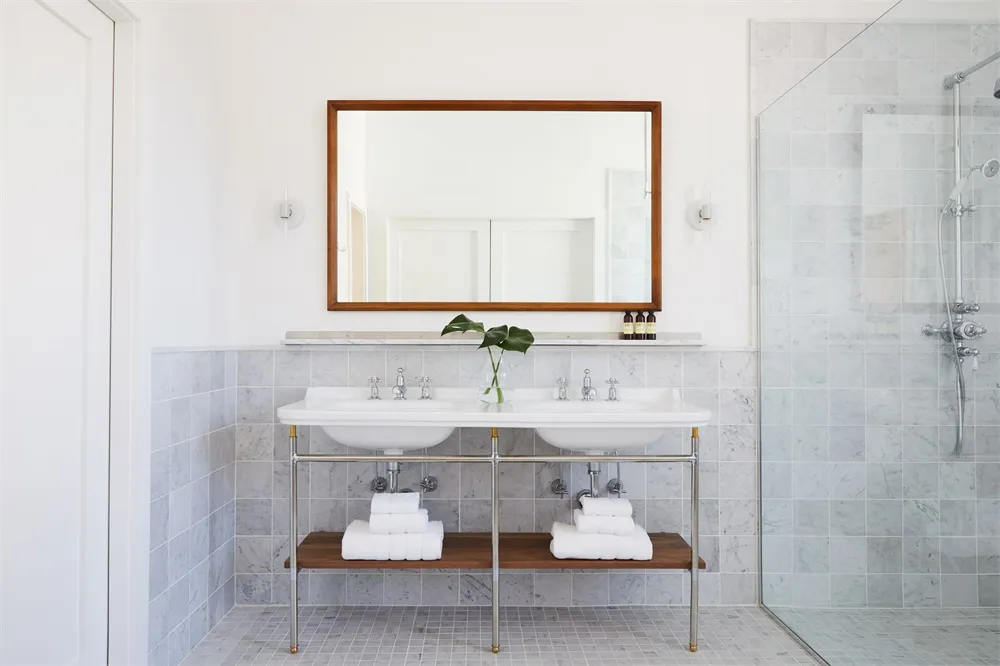 A modern bathroom with white marble tile walls, floor, and two white sinks with chrome faucets and a wooden shelf below. A gr