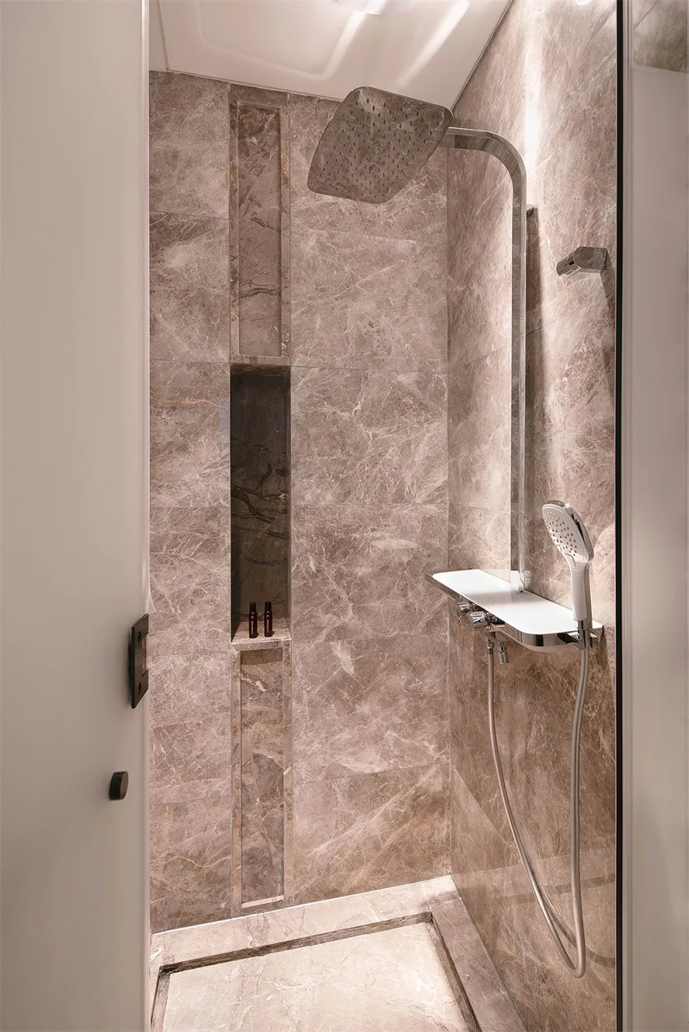 Modern bathroom with marble walls, shower head, and shelf with bottles.