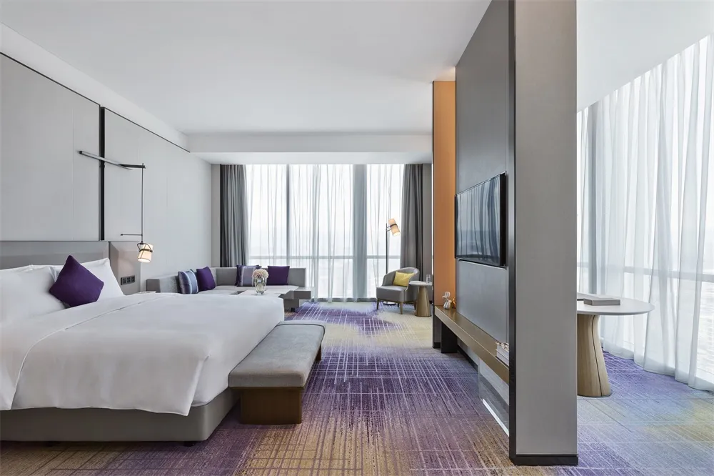 Modern hotel room with a large bed, sofa, television, and floor-to-ceiling windows.