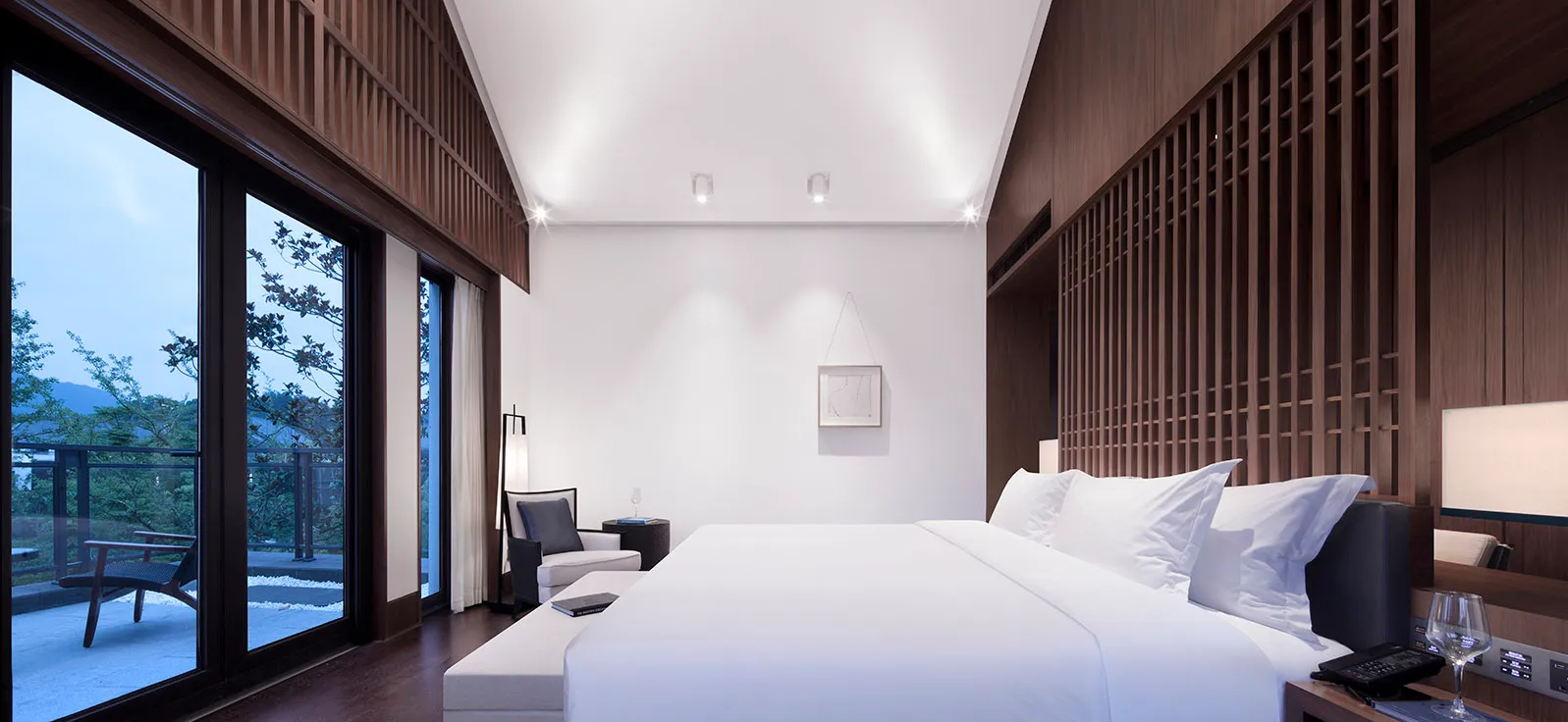 Minimalist-style bedroom featuring a large bed with a white sheet and dark wooden accents, with a view of the surrounding tre