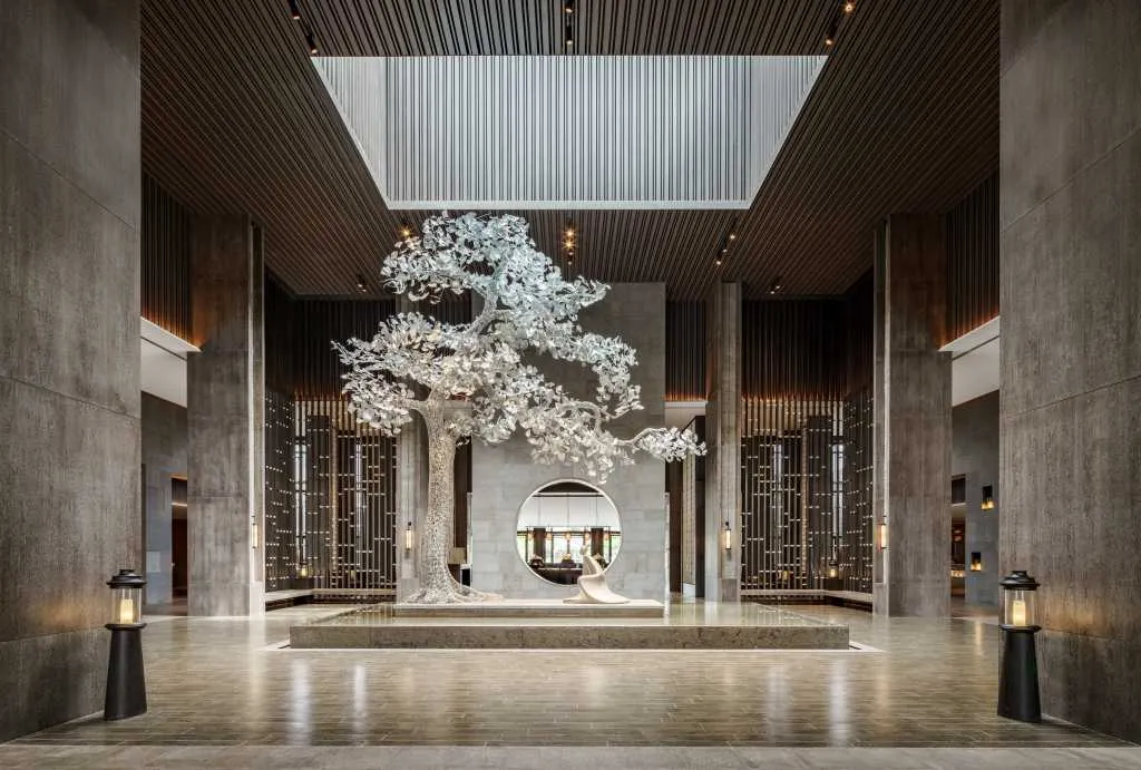 A modern hotel lobby featuring a large, metallic tree sculpture on a stone pedestal with tall columns and a skylight.