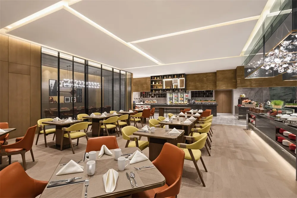 A spacious, modern restaurant interior with wooden tables, yellow chairs, and a well-lit kitchen area.
