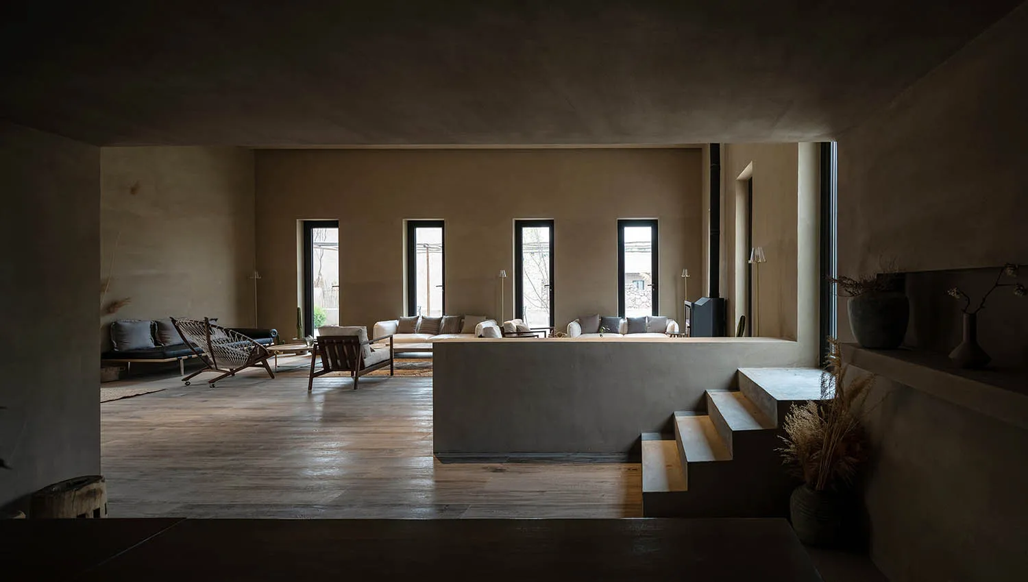 A minimalist living room featuring concrete walls and floors, with a central raised seating area and large windows.