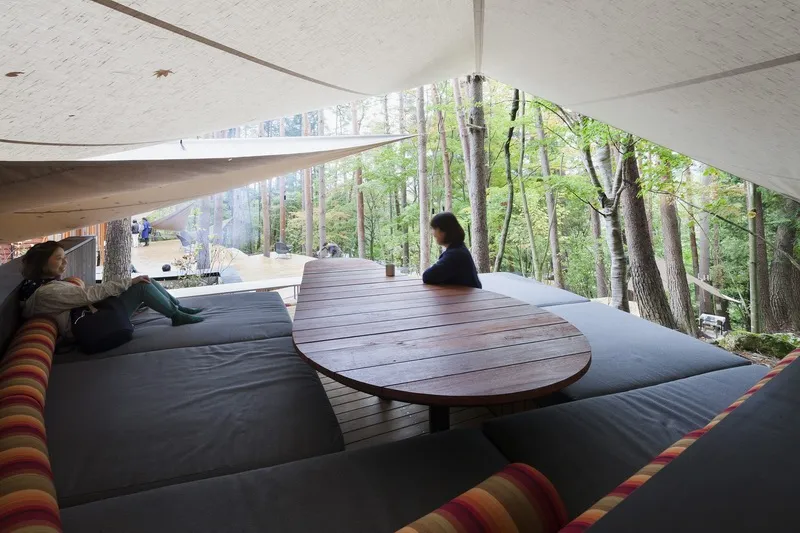 A modern, industrial-style lounge area nestled within a forest, featuring cozy sofas and a wooden table on a wooden floor, wi