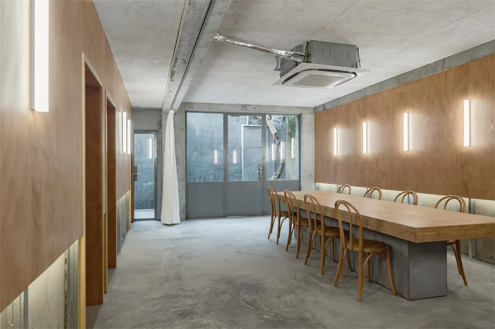 Industrial-style meeting room with concrete walls and floor, long wooden table with chairs, and a large window.
