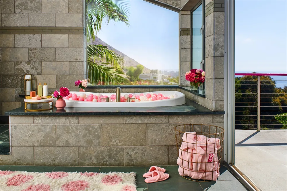 A modern spa bathroom with stone tile walls, a large window overlooking a mountain view, a bathtub filled with colorful balls