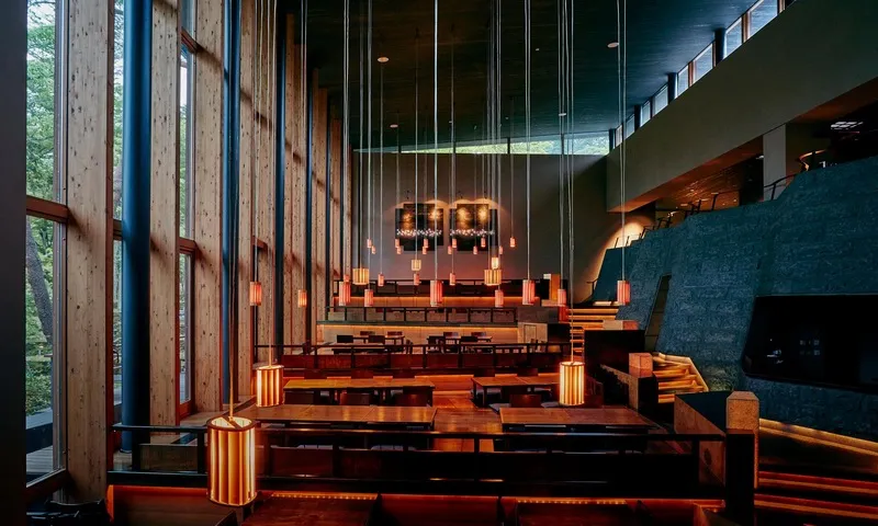 A modern restaurant interior featuring wooden tables and chairs, with hanging lights and a large window showing natural light