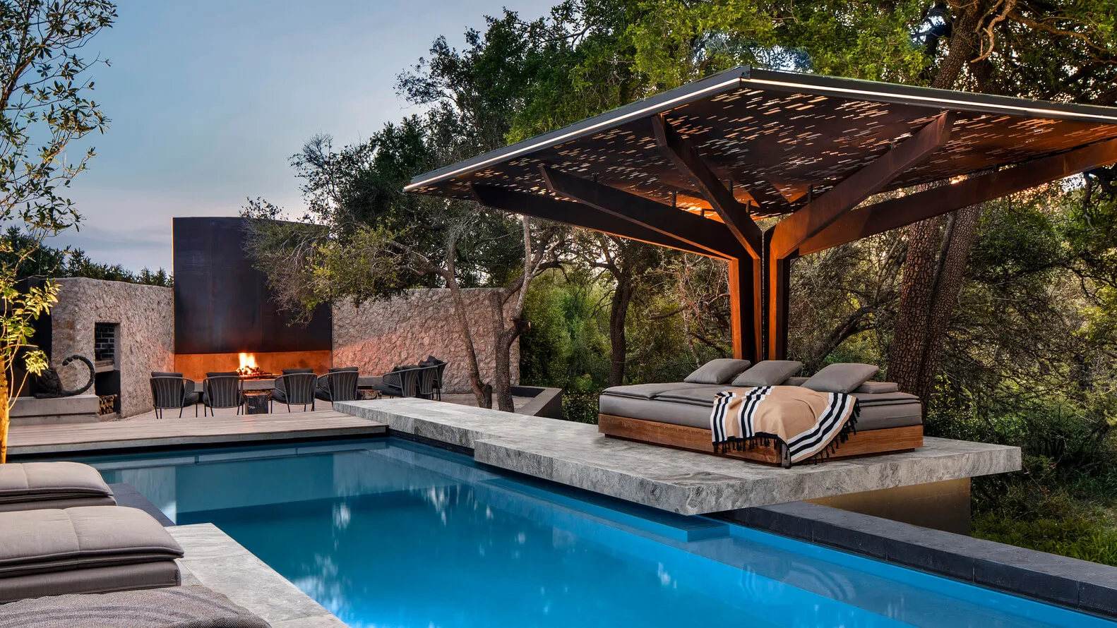 A modern residential pool side terrace featuring a fire pit, pergola, and seating area surrounded by natural stone and concre
