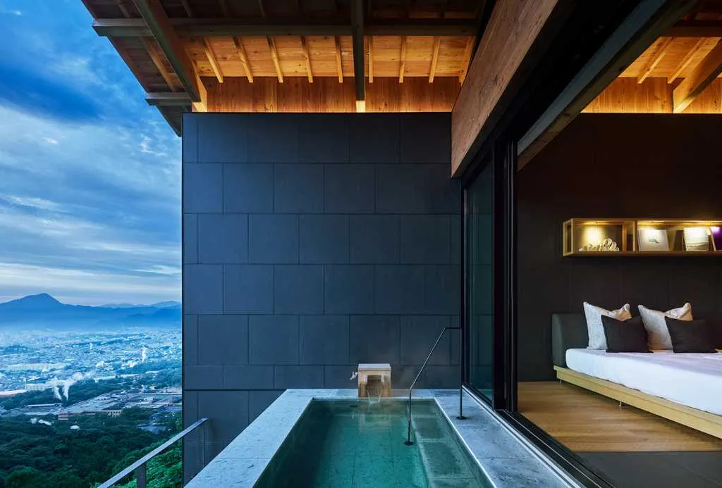 Modern bedroom featuring a black tiled wall, wooden paneled ceiling, and a green water feature overlooking a cityscape.