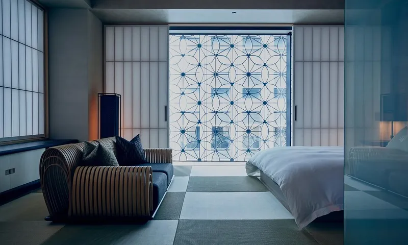 A minimalist hotel bedroom with a wooden sofa, a bed with white linens, and a sliding door with a geometric pattern.