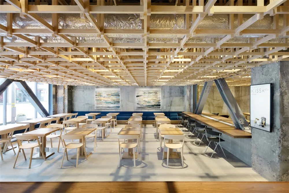 A modern cafe interior featuring wooden tables and chairs, exposed wooden beams, and concrete walls and flooring.