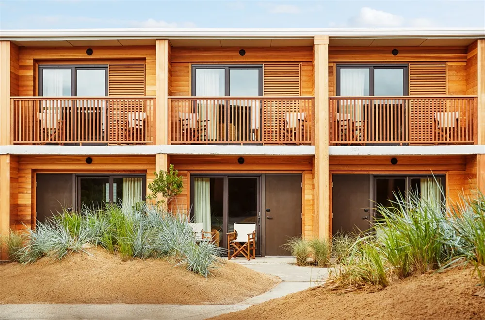 A two-story vacation home with a wooden exterior, outdoor seating areas, and sandy surroundings.
