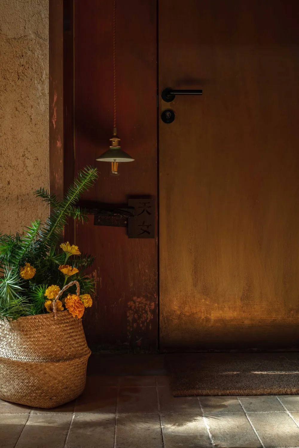 A wooden door adorned with a woven basket containing yellow flowers and greenery, illuminated by a hanging lamp.