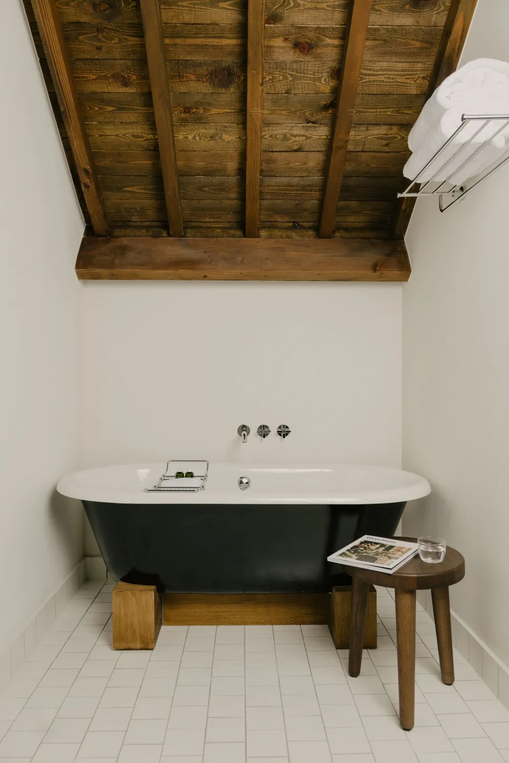 A minimalist bathroom featuring a wooden ceiling with beams, a white tub with black trim, and a wooden stool with a magazine